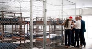 Four adults stand inside a fenced area with rows of metal bunk beds and thin mattresses, appearing to inspect the facility. The setting looks like a temporary or institutional shelter.
