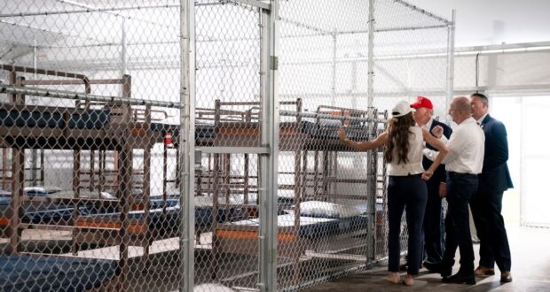 Four adults stand inside a fenced area with rows of metal bunk beds and thin mattresses, appearing to inspect the facility. The setting looks like a temporary or institutional shelter.