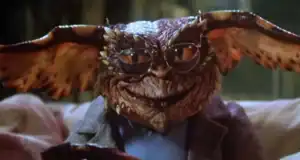 A gremlin creature with large ears, brown scaly skin, and glasses sits on a couch wearing a gray jacket, smirking at the camera.