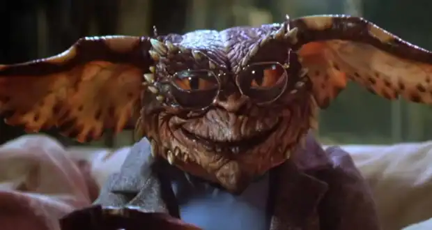 A gremlin creature with large ears, brown scaly skin, and glasses sits on a couch wearing a gray jacket, smirking at the camera.