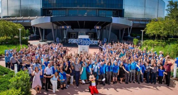 A large group of people, many wearing blue shirts, gather outside a building with "Test Track" signage. They are smiling, waving, and posing with a person dressed as Mickey Mouse in front. Lush greenery surrounds the area.