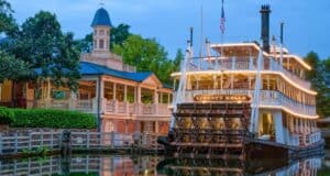 A white, paddlewheel riverboat with glowing lights, named “Liberty Belle,” is docked by a wooden riverside landing with a gazebo and lush trees in the background at dusk.