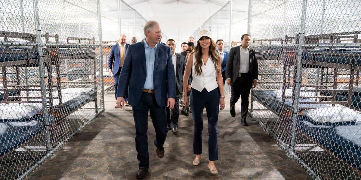 A group of people walk between rows of bunk beds inside a fenced facility with metal walls and floors, resembling a detention or shelter area. Two people lead the group, talking as they walk.