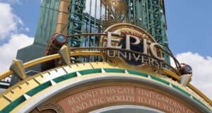 A close-up of the entrance sign for Universal Epic Universe, featuring gold, blue, and green decorative elements, with a blue sky and clouds in the background. Text below reads: "Beyond this gate find gardens and epic worlds to fill you.