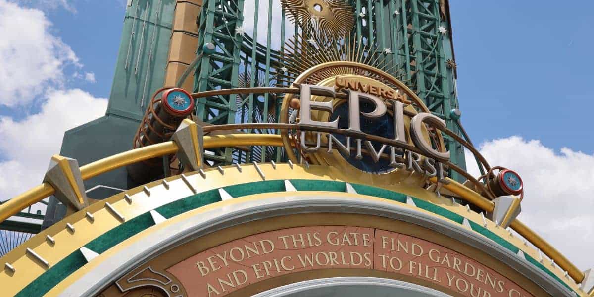 A close-up of the entrance sign for Universal Epic Universe, featuring gold, blue, and green decorative elements, with a blue sky and clouds in the background. Text below reads: "Beyond this gate find gardens and epic worlds to fill you.