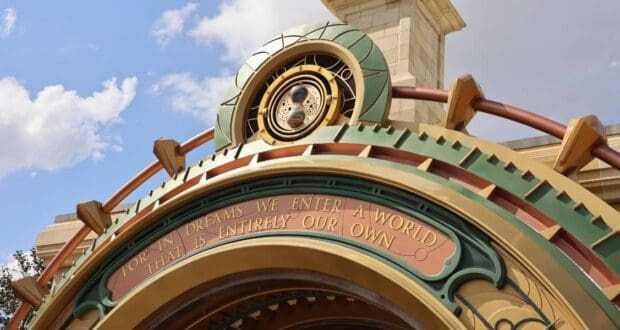 The image shows an ornate golden archway with a clock and gears above an engraved quote reading, “For in dreams we enter a world that is entirely our own,” against a blue sky with clouds.
