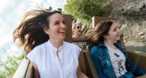 Three women ride a roller coaster outdoors, smiling and laughing as the wind blows their hair—a thrill much like Animal Kingdom attractions. The woman in front wears a white shirt, beside her is one in a denim jacket, and the third in back wears sunglasses.