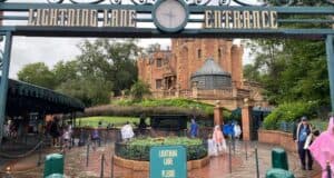 Entrance to the Haunted Mansion ride at a theme park, with a sign reading "Lightning Lane Entrance." Guests, some in rain ponchos, walk nearby on a cloudy day. The mansion’s gothic architecture is visible in the background.