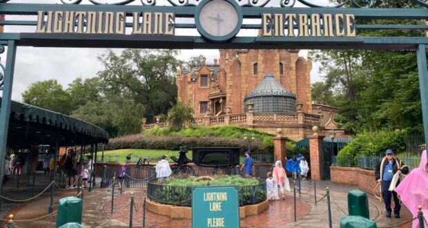 Entrance to the Haunted Mansion ride at a theme park, with a sign reading "Lightning Lane Entrance." Guests, some in rain ponchos, walk nearby on a cloudy day. The mansion’s gothic architecture is visible in the background.