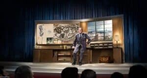 A man in a suit stands smiling in front of a large desk with maps and plans on display, set on a stage with blue curtains. An audience watches him from the foreground.