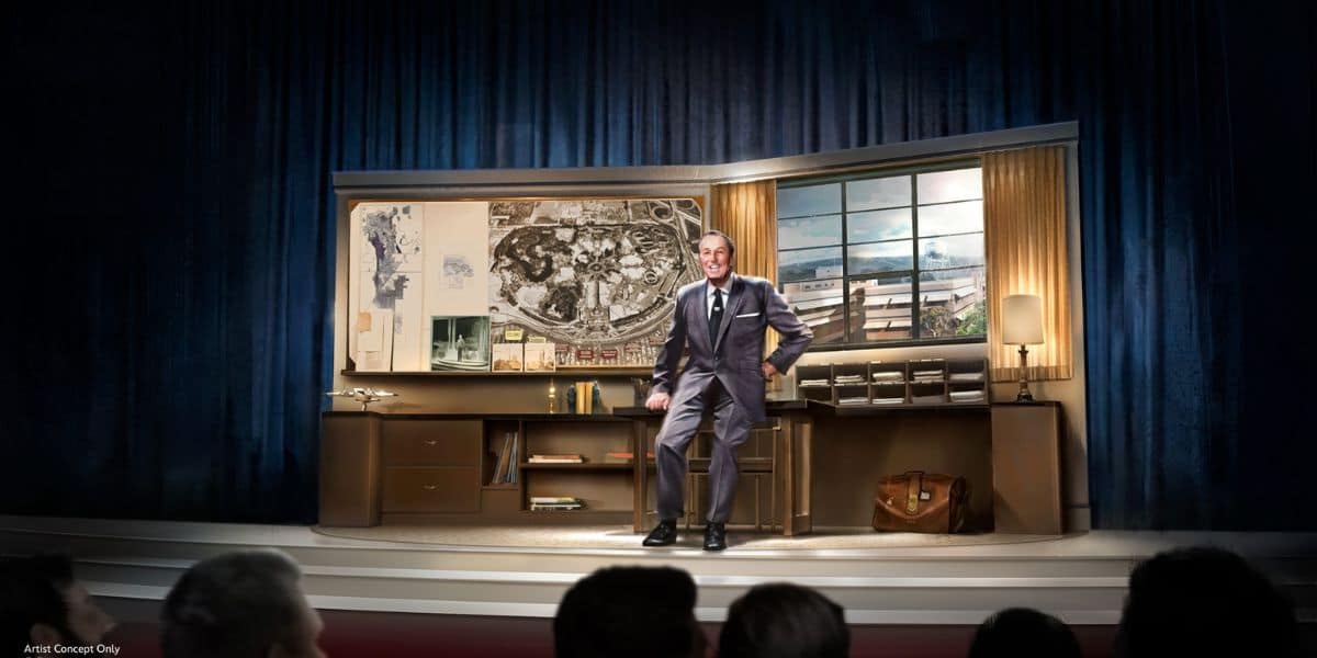 A man in a suit stands smiling in front of a large desk with maps and plans on display, set on a stage with blue curtains. An audience watches him from the foreground.