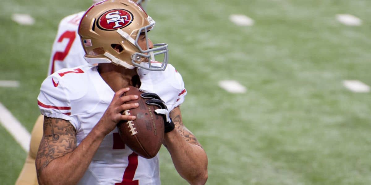A football player in a San Francisco 49ers uniform holds a football, preparing to throw during a game on a field. He is wearing a gold helmet and white jersey with red numbers.