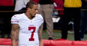 A football player wearing a white San Francisco 49ers uniform with the number 7 stands on the field, looking to the side. He has short hair and visible tattoos on both arms. The background shows blurred people and equipment.