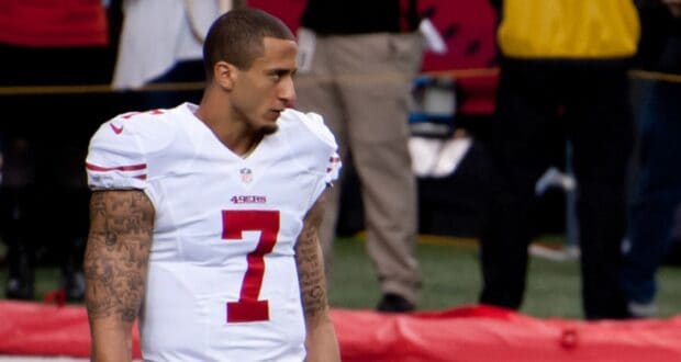 A football player wearing a white San Francisco 49ers uniform with the number 7 stands on the field, looking to the side. He has short hair and visible tattoos on both arms. The background shows blurred people and equipment.