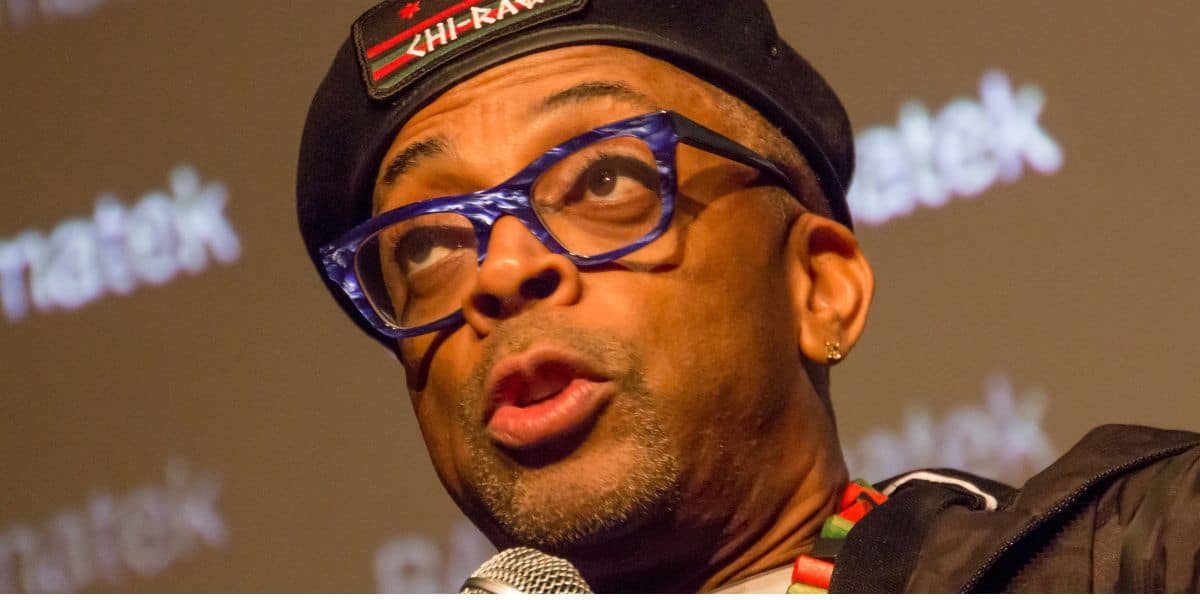 A man wearing blue glasses, a black beret with "CHI-RAQ" written on it, and a colorful necklace speaks into a microphone against a blurred background with white text.