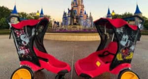 Two empty red and black strollaers, featuring images of Mickey and Minnie Mouse, are parked facing each other on an empty path in the Magic Kingdom. The strollers are positioned in front of a castle with blue rooftops and a statue, set against a clear sky in the amusement park.
