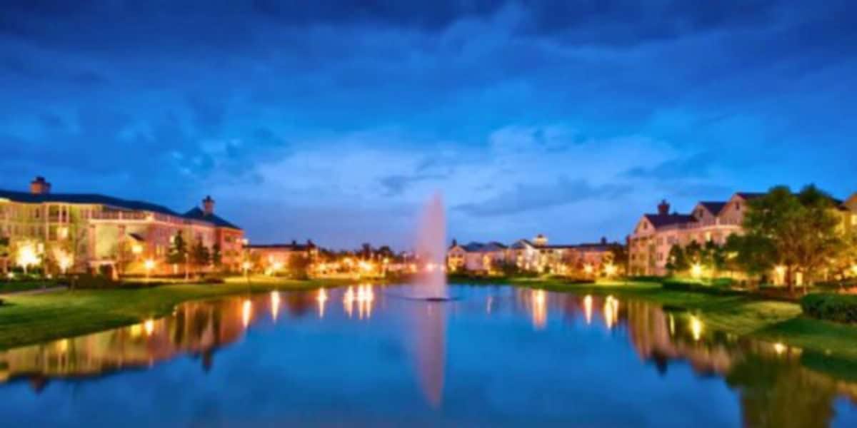 A serene evening view of a Disney World waterfront neighborhood with lighted buildings reflecting on the calm water. A central fountain gently sprays water upwards. The sky is a mix of deep blue and light clouds, creating a peaceful ambiance.