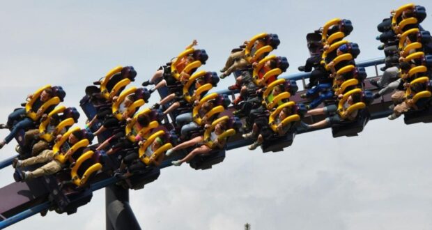 People ride a roller coaster with yellow harnesses, hanging upside down as the ride loops through the air against a cloudy sky. Many riders have their arms raised, expressing excitement and thrill.
