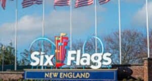 Entrance sign for Six Flags New England with colorful flags above the logo and American flags waving on flagpoles in the background under a partly cloudy sky.