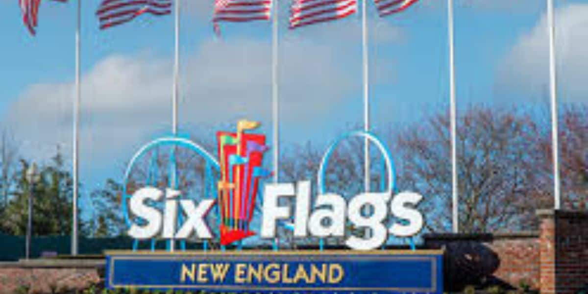 Entrance sign for Six Flags New England with colorful flags above the logo and American flags waving on flagpoles in the background under a partly cloudy sky.