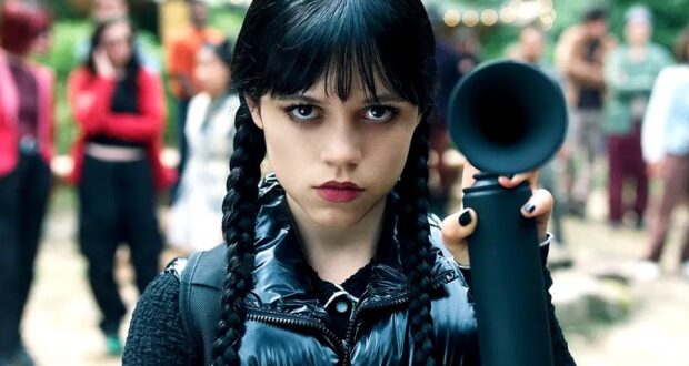 A girl with long black braided hair and dark clothing stares intensely at the camera while holding up a black object, with a blurred group of people standing in the background outdoors.