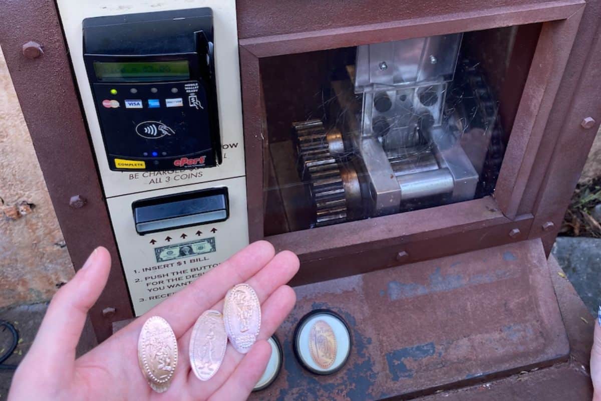 A hand holds three pressed souvenir pennies in front of a Disney World penny-press machine.