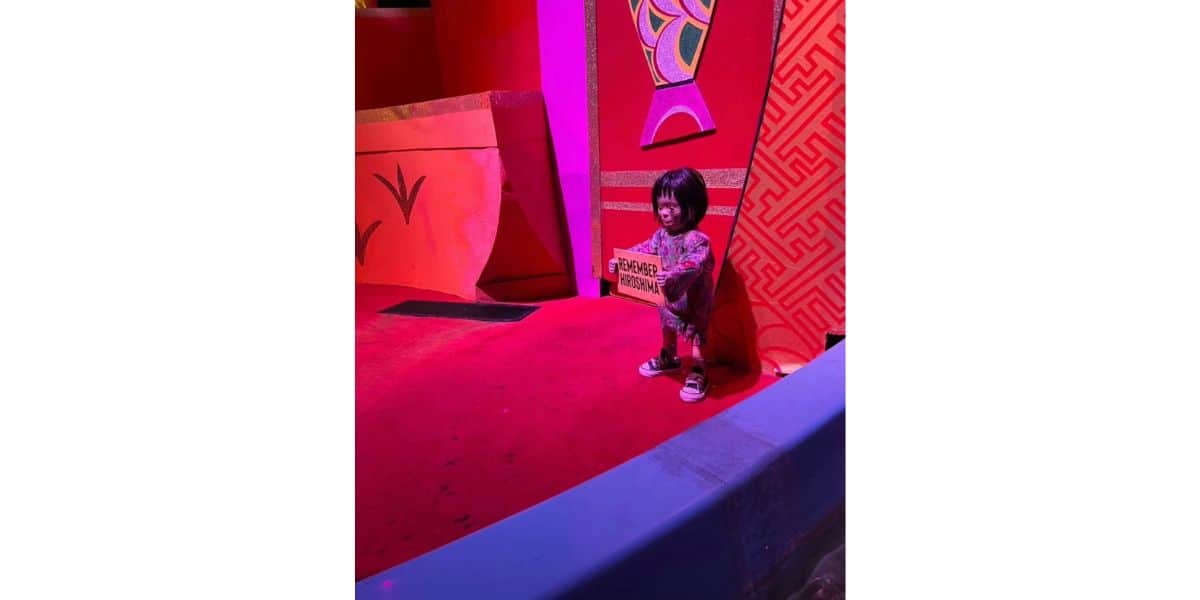 A young child stands on a red stage holding a rectangular sign with writing on it, surrounded by bright red and orange decorative walls and colorful patterns.