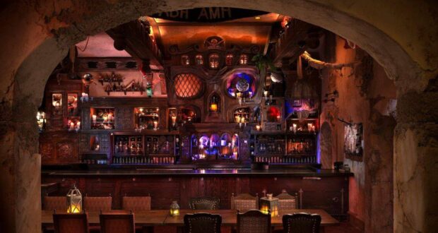 A dimly lit, intricately decorated bar with rustic wooden furniture, glowing lanterns, and shelves filled with bottles and curiosities, viewed through a stone archway.