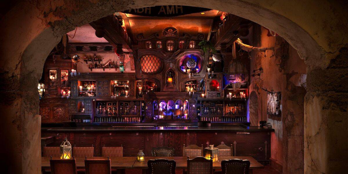 A dimly lit, intricately decorated bar with rustic wooden furniture, glowing lanterns, and shelves filled with bottles and curiosities, viewed through a stone archway.