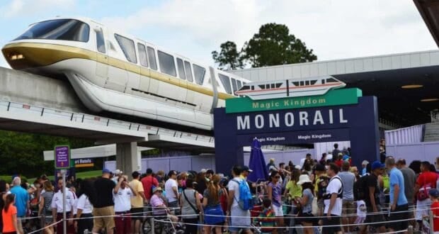 A large crowd waits beneath the "Magic Kingdom Monorail" sign as a monorail train arrives on the elevated track above the station on a sunny day.
