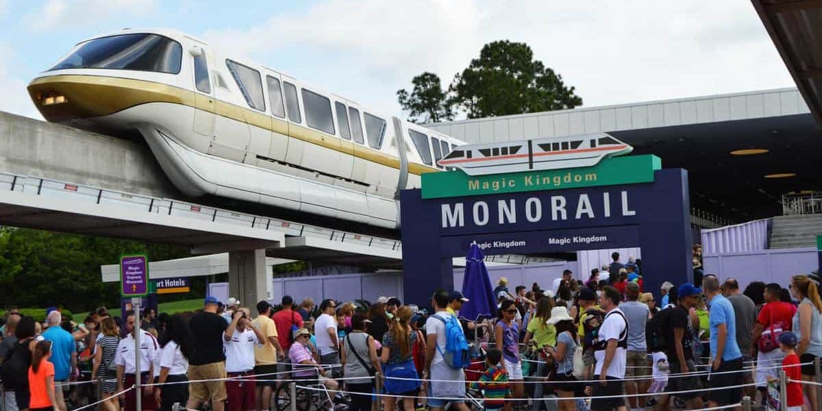 A large crowd waits beneath the "Magic Kingdom Monorail" sign as a monorail train arrives on the elevated track above the station on a sunny day.