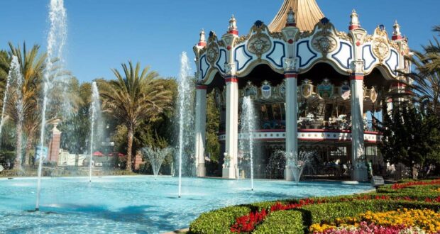 A colorful carousel with ornate decorations stands behind a large, clear fountain surrounded by palm trees, flowers, and neatly manicured greenery under a bright blue sky.
