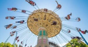 People ride a large swing carousel at an amusement park, with chairs spinning outward under a clear blue sky. The ride’s ornate top and tall central column are visible, surrounded by trees and roller coaster tracks.