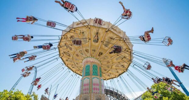 People ride a large swing carousel at an amusement park, with chairs spinning outward under a clear blue sky. The ride’s ornate top and tall central column are visible, surrounded by trees and roller coaster tracks.