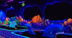 A glow-in-the-dark mini-golf course with an underwater theme, featuring colorful sea creatures, coral, and a large whale sculpture under black lights. Neon colors and sea decorations line the walls and course.