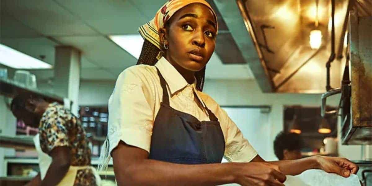 A woman wearing a patterned headscarf and apron works in a restaurant kitchen, looking to the side. Two other people are visible in the background, also busy with their tasks.