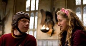 A young man in a brown Quidditch helmet and red sweater looks at a smiling young woman with long curly hair and a pink headband, sitting together in a medieval-style hall with stone walls and arched windows.