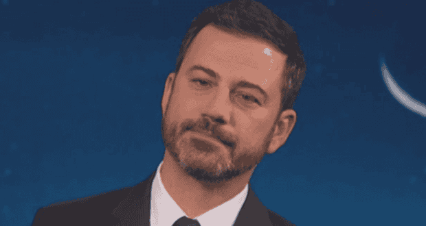 A man with short dark hair and a beard, wearing a suit and tie, stands against a dark blue background with a crescent moon visible. He has a serious, slightly skeptical expression.