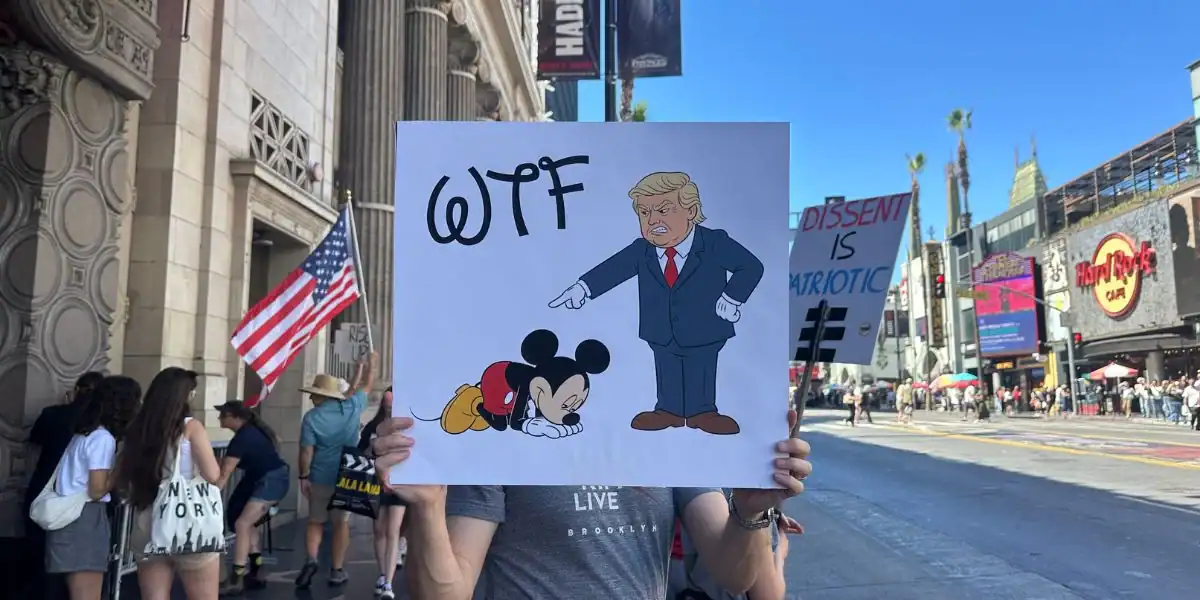 A person holds a sign showing a cartoon of Donald Trump pointing at a sad, kneeling Mickey Mouse with "WTF" above them. People and American flags are visible on a city street in the background.