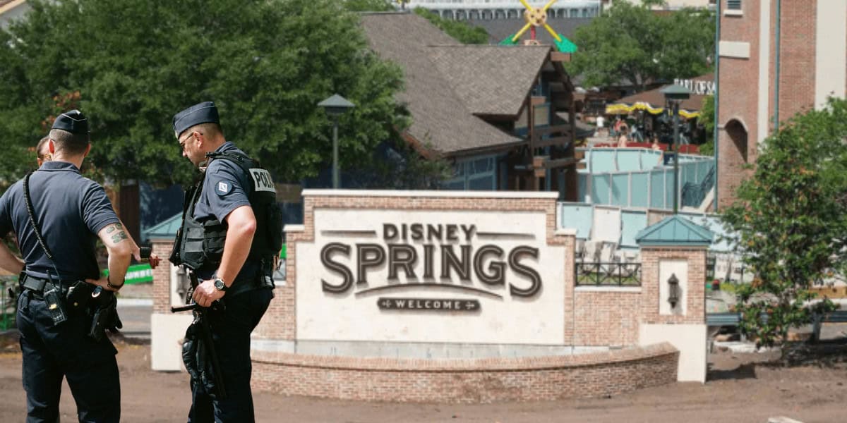 Two police officers in uniform stand in front of the Disney Springs welcome sign, with buildings and trees visible in the background on a sunny day.