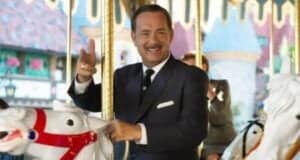 A man in a suit smiles and waves while riding a white carousel horse. The background features colorful decorations.