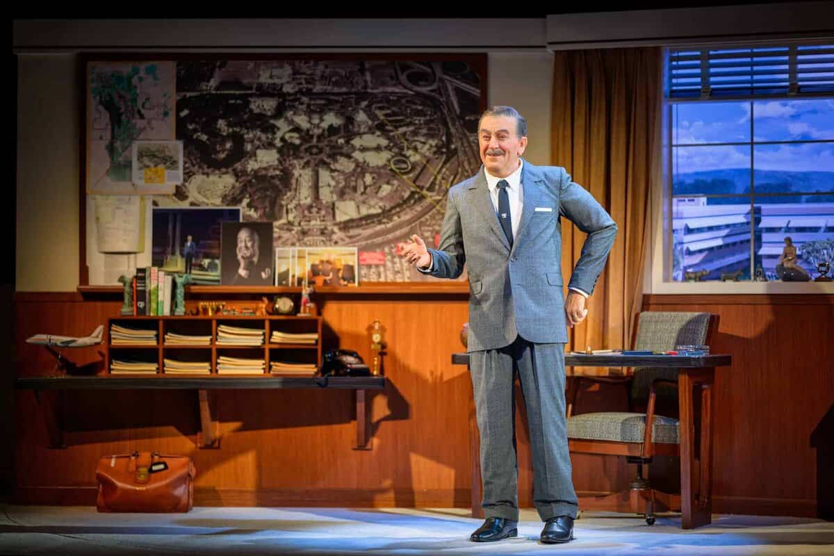 A man in a gray suit stands smiling in a vintage office with a desk, shelves, and a map-covered wall. Sunlight streams through a window, revealing mountains outside. The scene appears to be from a stage play.