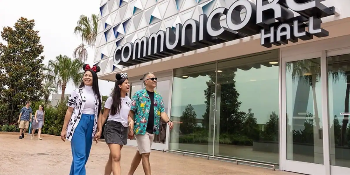 Three people, two women with Disney ears and a man in a tropical shirt, walk hand-in-hand outside CommuniCore Hall, a modern building with large glass windows and geometric patterns.