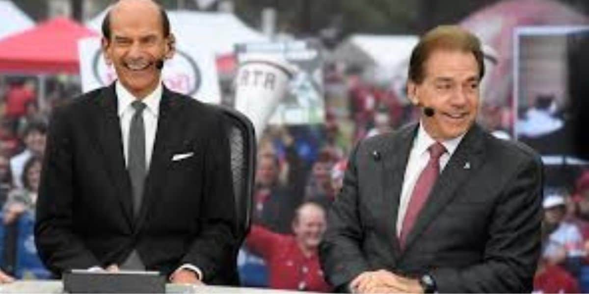 Two men in suits sit at a desk outdoors, smiling during a broadcast. A crowd with red and white attire and signs is visible in the blurred background.