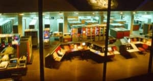 A large, brightly lit vintage computer control room with multiple rows of mainframe computers, control panels, and monitors, viewed from above through a window.
