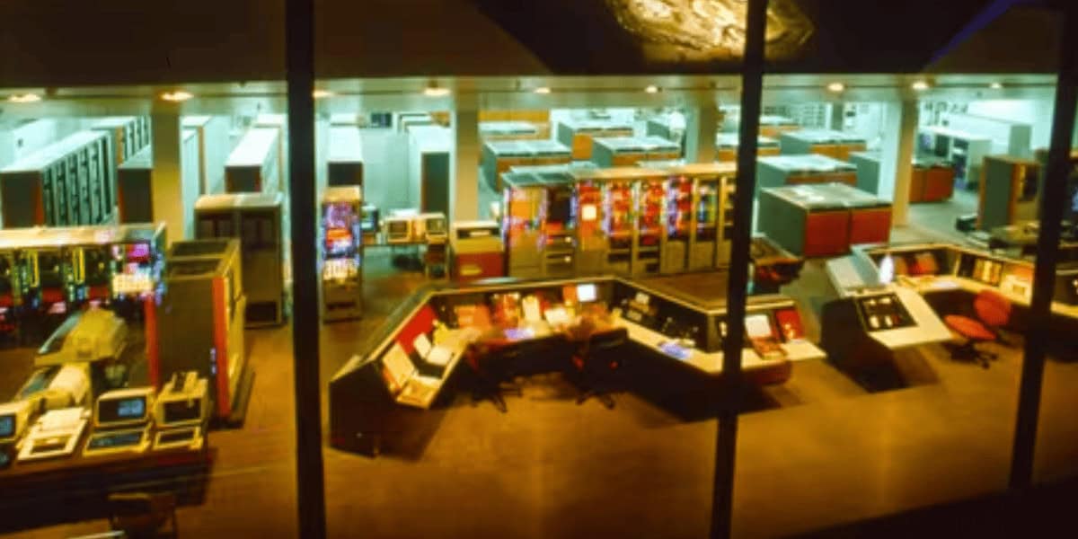 A large, brightly lit vintage computer control room with multiple rows of mainframe computers, control panels, and monitors, viewed from above through a window.