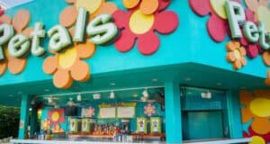 A colorful outdoor bar called "Petals" features bright flower decorations and a turquoise exterior, with a counter displaying drinks and menus, evoking a playful, retro vibe.