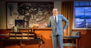 A man in a gray suit stands and gestures in a vintage office with maps, photos, and awards on the wall behind him. A desk, chair, and window with a blue sky are also visible in the room.