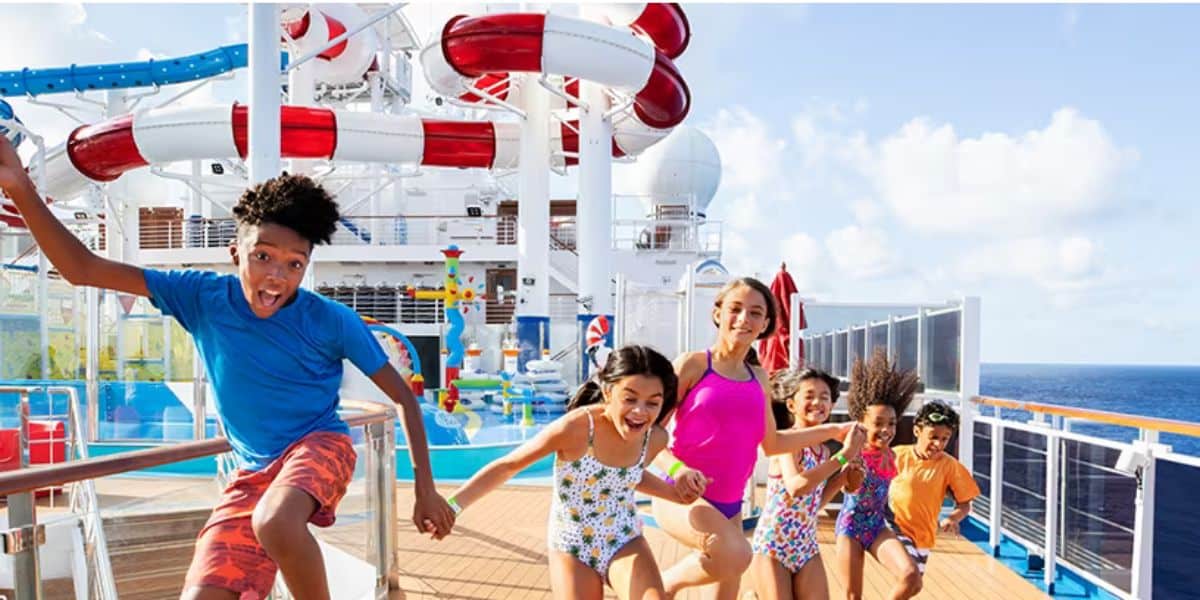 A group of smiling children and one adult in swimsuits run excitedly on a cruise ship deck near a large water park with slides, under a blue sky.
