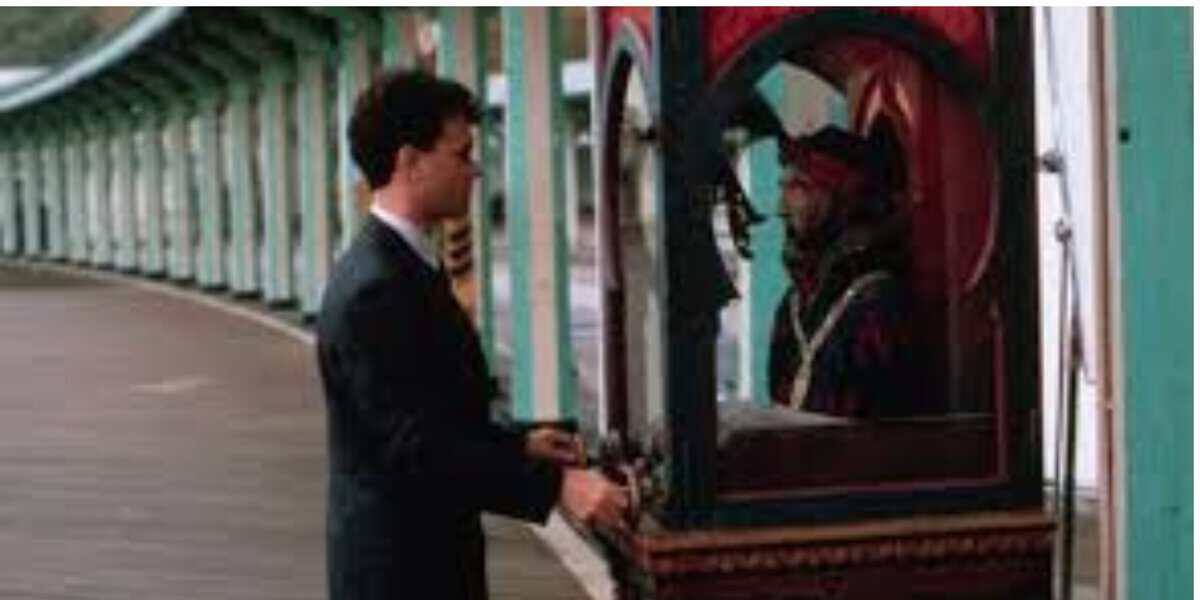 A man in a suit stands in front of a fortune teller machine on a boardwalk, holding something in his hand, with a row of green supports in the background.